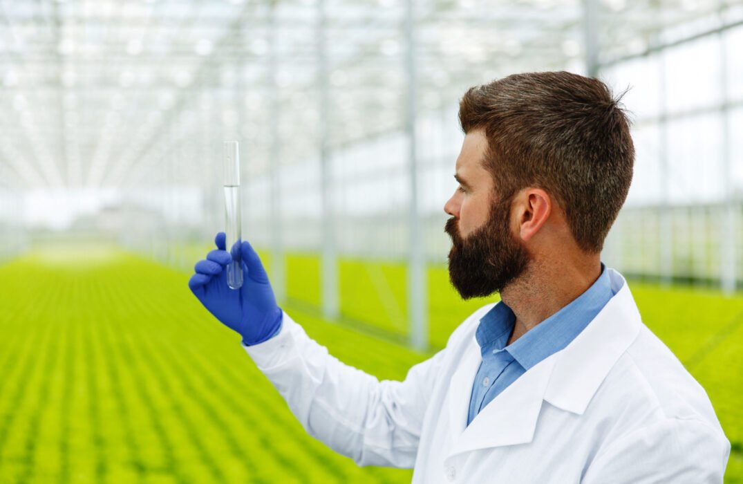 Thoughtful man researcher holds a glass tube with sample standin Thoughtful man researcher holds a glass tube with sample standing before plants in the greenhouse