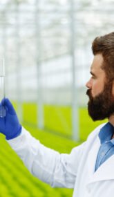 Thoughtful man researcher holds a glass tube with sample standin Thoughtful man researcher holds a glass tube with sample standing before plants in the greenhouse